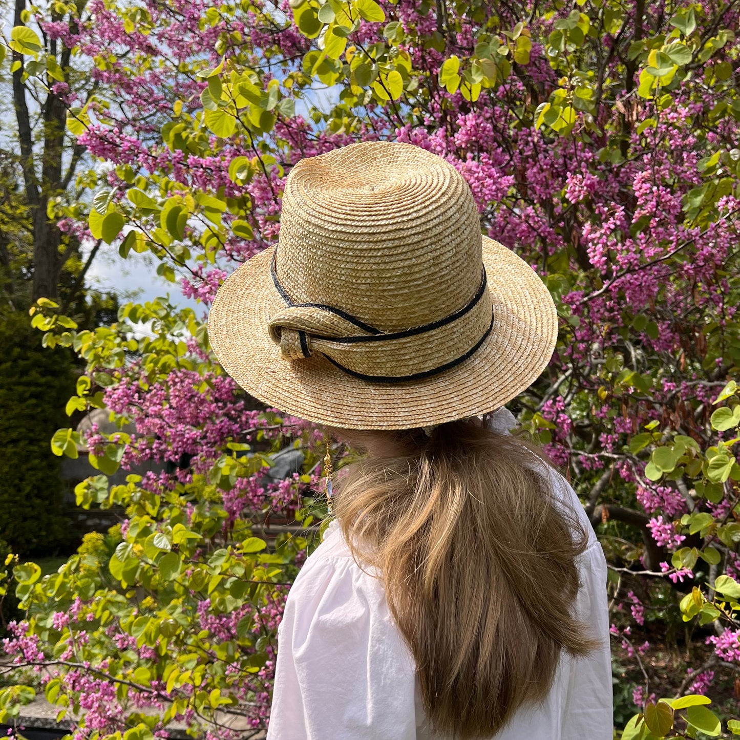 Person wearing a straw hat and white coat standing in front of purple flowering trees.