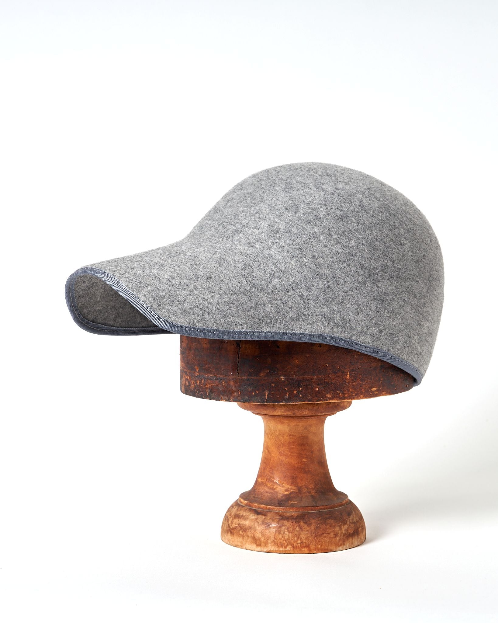Grey felt TO’CA Shell cap on a wooden stand against a white background