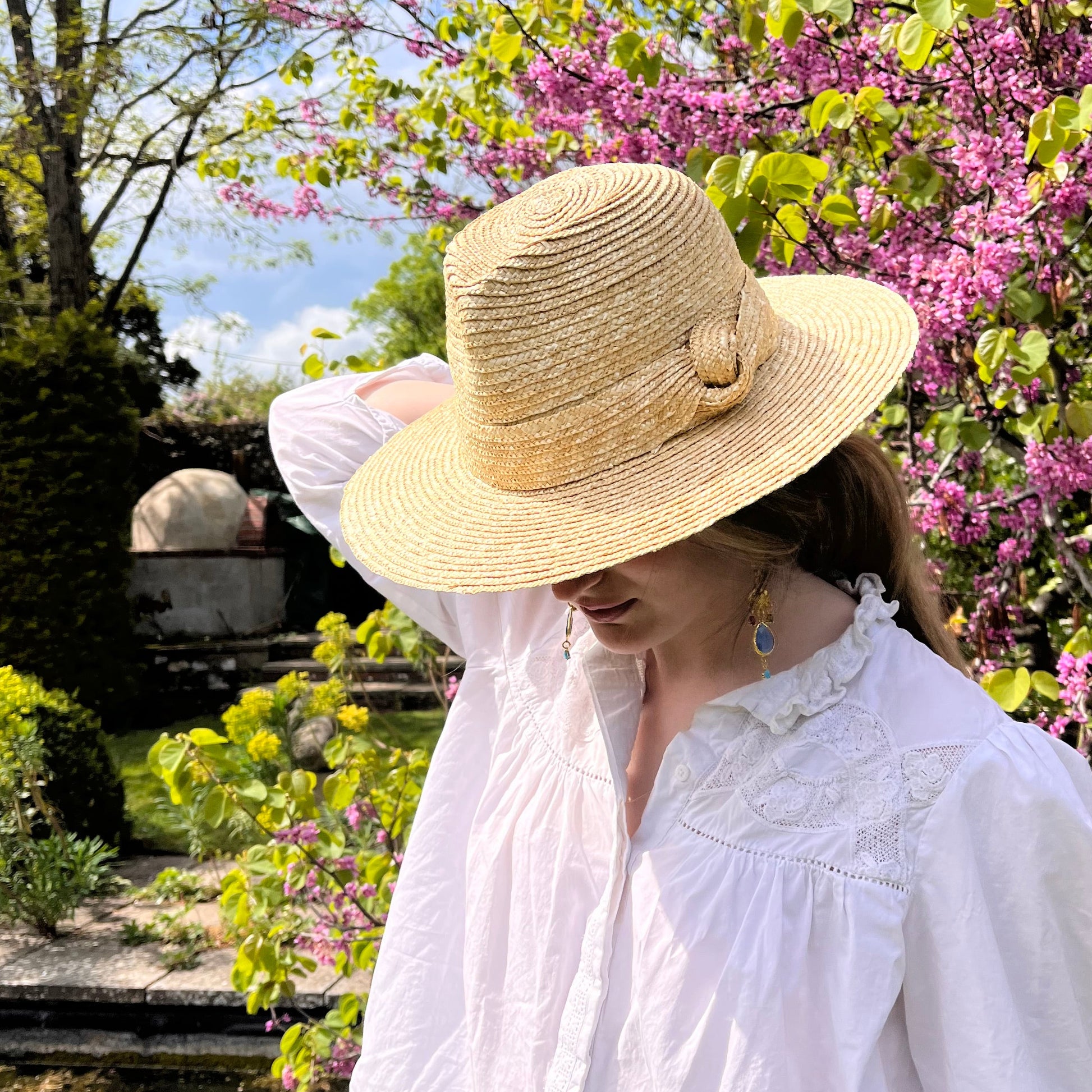 Person wearing a straw hat and white dress standing in front of pink flowering trees.