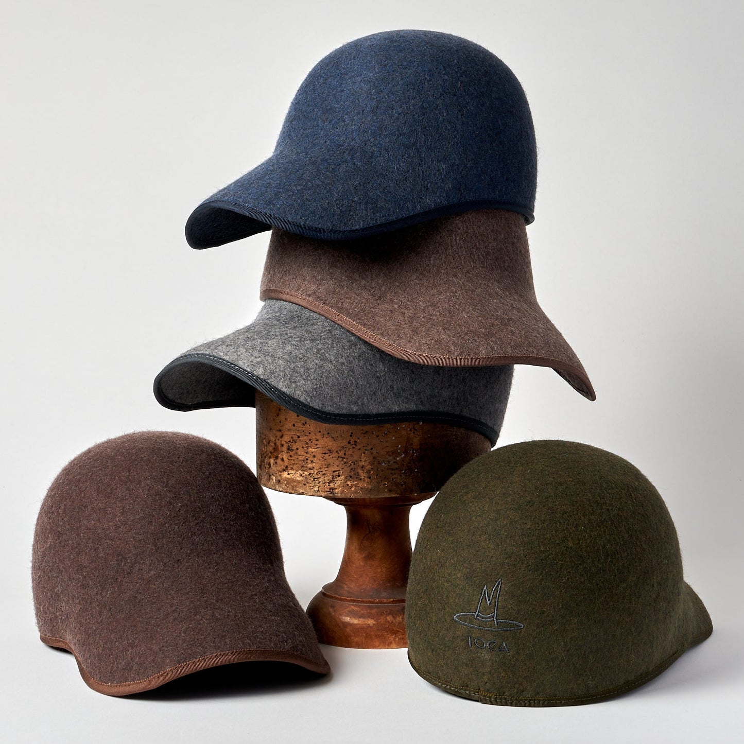 Four felt hats in different colors stacked on a wooden stand against a light gray background.