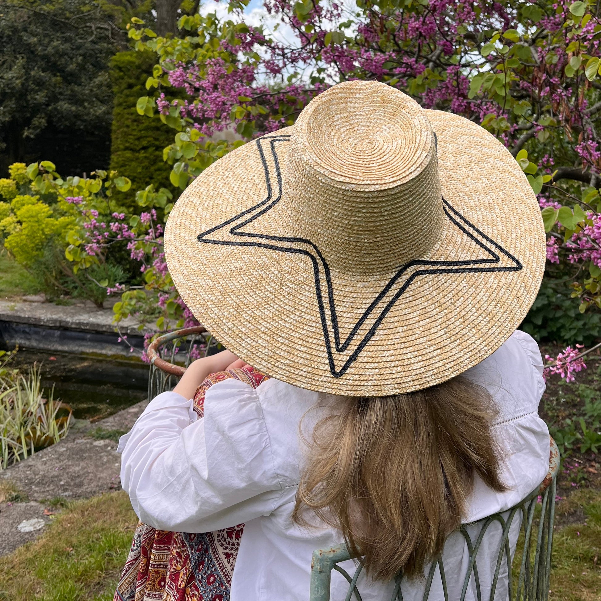 Person wearing a straw hat with a star design, standing in a garden with pink flowers and greenery.